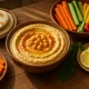 Bowl of classic hummus with olive oil and paprika, surrounded by fresh vegetables and pita on a rustic table