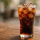 Refreshing glass of Coca-Cola with ice cubes and fizz on a wooden table, showing the look of a sweet carbonated drink in summer light.