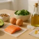 Nutrient-rich foods for postpartum hormone balance including avocado, salmon, eggs, nuts, spinach, quinoa, olive oil, and supplements arranged on a clean kitchen table in natural daylight.
