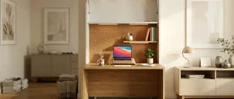 A horizontal view of a cozy and functional home office integrated into a white wall cabinet. The desk is open, revealing a warm wooden interior with a laptop, a steaming mug, and a small plant. Soft warm lighting illuminates the workspace, while the rest of the room is bright and minimalist with neutral decor.