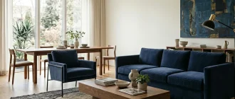 Modern living room interior featuring a timeless navy blue velvet sofa, a matching armchair with metal legs, and a solid wood coffee table against a neutral background.