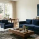 Modern living room interior featuring a timeless navy blue velvet sofa, a matching armchair with metal legs, and a solid wood coffee table against a neutral background.